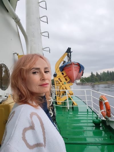 A medium shot portrait features a woman with peachy-pink hair posing on the deck of a vessel. She is wearing a white sweater with large, light brown heart patterns across the back and shoulders. The woman is looking at the camera with a slight smile and has on dark eyeliner and pink lipstick. Behind her, a bright orange life preserver is attached to a white railing. The green deck is wet and reflects the overcast sky. In the background, a yellow crane holds a red lifeboat, and a body of water is visible, bordered by a treeline. The scene appears to be on a river or canal. The location is not specified as Belousovo, Russia, and there is no evidence in the image to support this claim.