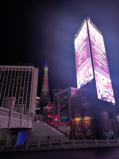 The image captures a nighttime view of the Las Vegas Strip, featuring a prominent, illuminated tower advertising the Horseshoe casino and hotel. The tower's digital billboard displays a vibrant pink hue and advertises an "ULTIMATE STEAKHOUSE TOUR" with details about restaurants, meals, and pricing, along with a call to action to book at Caesars.com. To the left of the Horseshoe tower, the Eiffel Tower replica of Paris Las Vegas is visible, lit up in red, white, and blue. A tall, modern hotel building, likely part of the Paris Las Vegas property, stands to the far left, its windows lit from within. In the foreground, a pedestrian walkway with an escalator leads up towards the casino entrance, with several individuals ascending. The environment is urban, with streets and sidewalks visible in the lower portion of the image, indicating a public space in a bustling city. The overall scene is characterized by bright neon lights and architectural structures, typical of the Las Vegas entertainment district.