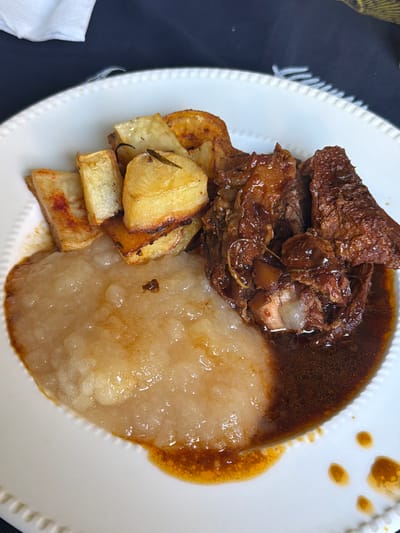A close-up shot of a white plate featuring a meal. The meal comprises of a serving of roasted potatoes, braised meat in a dark brown sauce, and what appears to be a soft, light-colored purée or mash, potentially root vegetables. The plate has a decorative edge, with visible details of the dish filling the frame. The overall environment, including the location of La Lucila, Argentina, cannot be determined from the image itself.