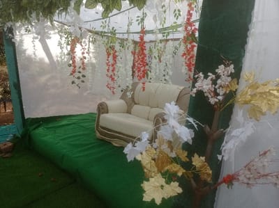 The image displays a decorated outdoor event setup in Ugbongha, Nigeria. A light beige two-seater sofa with tan trim is centrally positioned on a raised platform covered in green artificial turf. Overhead, a canopy structure is adorned with hanging artificial white and orange-red wisteria-like flower garlands and green foliage. The back and left sides of the canopy are enclosed by translucent white fabric, while a vertical support on the right is covered in green fabric. In the foreground to the right, a decorative arrangement of artificial branches features white and gold maple-like leaves and small white blossoms. The blurred background, visible beyond the translucent fabric on the left, indicates an outdoor environment with trees. No individuals are present. A brick and dark material are visible on the ground to the far left, outside the elevated green platform.