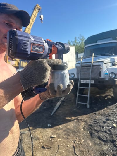 The image shows a person, possibly a man, wearing a baseball cap and work gloves, operating a blue and orange power drill. The drill has visible text including "MPSG550/800V", "220-240V-50Hz 500W", and "CEERE". The person is holding a cylindrical object, possibly a component or a piece of equipment, with the drill.

In the background, there is a large white vehicle, possibly a truck or an industrial vehicle, with a ladder attached to its side. License plate text "P93603 112" is visible on the vehicle. Further in the background, a yellow construction crane is visible against a clear blue sky, suggesting an outdoor industrial or construction setting. The ground appears to be rough and sandy, with some debris.

The lighting indicates it is daytime, likely sunny. The overall scene suggests a work or repair activity is in progress, possibly in an outdoor industrial area in Parma, Russia, given the Russian license plate. The focus is on the person's hands and the tool, highlighting the act of using the power drill.