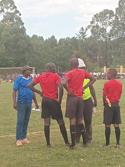 The image depicts an outdoor scene on a grassy field in Chevaywa ward, Kenya. In the foreground, five adult males are present. On the far left, a man wears a blue short-sleeved shirt, blue jeans, light brown shoes, and a camouflage print hat; he holds a white paper in his left hand and faces right. To his right, three men are dressed in matching red short-sleeved shirts, black athletic shorts, black knee-high socks, and athletic shoes, standing with their backs partially or fully to the viewer. Two of these individuals have their hands on their hips. The man on the far right has a yellow and red striped flag tucked into the waistband of his shorts. Partially visible between two of the men in red is a fifth individual wearing a lime green shirt and dark pants, holding a clear plastic bottle. In the midground, a white-netted soccer goal is visible on the left side of the field. A crowd of people is gathered in the distance across the field, partially obscured by a light-colored fence or wall on the right. The background consists of a dense line of green trees under an overcast sky.