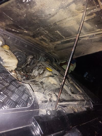 The image displays the open engine compartment of a black vehicle, likely a utility or off-road model, situated in Msata, Tanzania. The vehicle's hood is propped open by a metallic rod, revealing its dirty, textured underside with some paint wear. Within the engine bay, various components are visible, including a translucent fluid reservoir with a yellow cap on the left, and another engine part with a yellow cap deeper within the bay. The engine area is significantly soiled with dust and grime. A section of a black, diamond-plated fender or wing is visible on the left side, indicating the vehicle's robust construction. In the dimly lit background, to the right of the engine bay, a partial view of a person wearing a light-colored long-sleeved garment is discernable, suggesting human presence near the vehicle, possibly during maintenance or inspection. The overall scene indicates the vehicle is in a working condition, possibly under repair or inspection, in a dusty environment.
