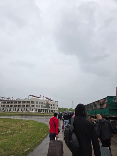This outdoor image, presented sideways, depicts a queue of people in what appears to be a transit or checkpoint area on an overcast and damp day. The sky is a uniform, heavy gray, and the ground is visibly wet from rain.

In the foreground, a person wearing a red hooded jacket and dark pants is seen from behind, pulling a dark roller suitcase. Ahead of them, a line of individuals stretches into the distance, many carrying luggage and some holding umbrellas to shield from the rain. Further along the queue, a person in a black hooded jacket carries a backpack, and closer to a large green truck, another individual with a bald head is seen in a black jacket. The activity suggests people are waiting to board a vehicle or pass through a control point, likely for travel.

To the right (if rotated), a large green truck or container forms part of the immediate background, alongside which the queue appears to be formed. Across a paved area and a strip of green grass, a multi-story, modern building stands. It has a light-colored facade with numerous windows and distinctive red architectural features, including ornate elements along its roofline and balconies. A logo or Chinese characters are faintly visible on the building's facade, consistent with the location context of Unknown, China. A red flag on a flagpole is also visible in the mid-ground. The overall scene conveys a sense of waiting and travel under gray, wet conditions.