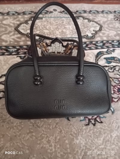 A black leather handbag with a textured finish is positioned centrally on a patterned rug. The handbag has two rounded handles and a prominent embossed logo featuring the letters "miumiu". The rug has a light beige base with intricate patterns in muted colors, including shades of gray, blue, and pink. The lighting is soft, casting subtle shadows. The timestamp "30.07.2026 15:23" is visible in the bottom right corner of the image, and "POCO C65" is in the bottom left. This image was taken in Beruniy, Uzbekistan.