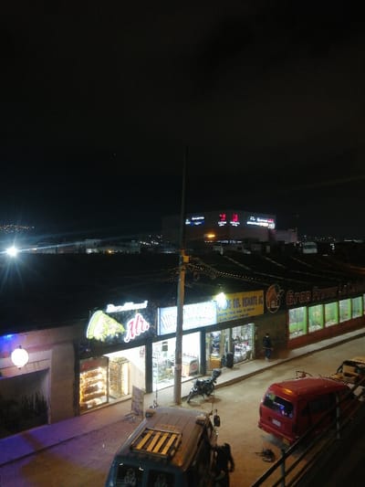 A nighttime street view featuring a row of businesses. "Mr." with a pizza slice graphic is visible, along with "Los Del Renate KC" and "Gran Fraver." The street is illuminated, showcasing parked motorcycles and vans. A large building with illuminated signs stands in the background on a higher elevation. The night sky is dark and clear.