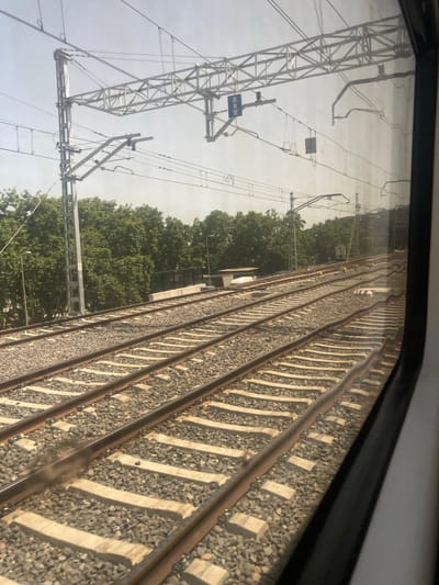 The image shows a view from a train window, looking out at multiple sets of train tracks. The tracks are ballasted with gravel and have concrete sleepers. Overhead, there are power lines supported by a metallic lattice structure and poles. Trees line the right side of the tracks, suggesting an urban or suburban environment. In the distance, a hill is visible. The sky is bright and hazy, indicating daytime. A blue traffic sign with a white symbol is visible on a pole near the overhead wires. Reflections are visible in the train window, obscuring some details. The image appears to be taken in Montcada i Reixac, Spain, a location known for its extensive railway infrastructure.