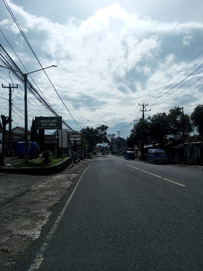 The image depicts a wide-angle view of a paved road extending into the distance under a bright, partially cloudy sky in the city of Domphoog_Dungkarchhoeling, Bhutan. The scene is static, showing no active movement or visible human figures.
The foreground is dominated by a dark asphalt road, marked with a solid white line on the left separating it from a paved sidewalk, and a solid yellow line further right along the road's center or shoulder. The sidewalk, constructed of paving stones and concrete, appears worn. A low, dark masonry wall separates the sidewalk from a narrow, grass-covered median strip with some small green plants.
Along the left side of the road, multiple utility poles are visible, heavily laden with a dense network of black overhead electrical and communication cables that converge towards the horizon. A street lamp is affixed to one of these poles. Behind the poles and foliage, a prominent dark rectangular sign with white text reads "MASJID AL-UM". Another sign with the word "KHITANAN" is partially visible to its right. Structures and buildings with varying rooflines, including one with a red-tiled roof, are partially visible behind the signs and utility infrastructure. A blue tarp-covered object is situated near these structures on the left.
The right side of the road features more utility poles with fewer visible overhead lines and numerous trees with dense green foliage. Buildings with dark or corrugated roofs are partially visible behind the trees and poles. Two blue minivans or small buses are parked along the curb on the right, facing away from the viewer, with the rear vehicle partially obscured by trees. Further down the road, a light-colored truck or similar vehicle is faintly discernible.
The overall environment is an urban streetscape, characterized by overhead utility lines, roadside vegetation, and commercial or residential structures. The lighting suggests daytime conditions with diffuse sunlight breaking through the clouds.