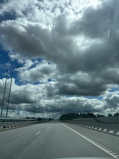 The image shows an aerial view of a highway in Lehmja, Estonia, with a cloudy sky and trees in the background. The highway is empty, with no cars or people visible. The sky is filled with dramatic, dark clouds that cast shadows on the road. The clouds are so dense that they obscure most of the sky, with only patches of blue visible. The trees in the background are lush and green, suggesting it is either spring or summer. The highway appears to be well-maintained, with clear lane markings and guardrails on either side. Streetlights are visible along the side of the highway.