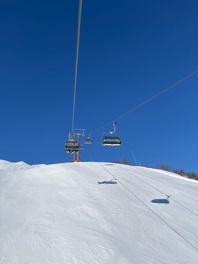 The image depicts a ski slope in Zürs, Austria, under a clear, deep blue sky. A multi-cabin chairlift system is ascending the snow-covered mountain. A main steel haul rope extends from the upper frame to the mid-ground. Multiple chairlift cabins are visible: one in the mid-left contains at least two passengers, while another in the mid-right is empty. A metallic grey chairlift pylon with an orange base stands on the slope, supporting the cables. The expansive white snow features visible ski tracks and grooming patterns. Elongated, dark shadows of the chairlift cabins and cables are cast onto the snow, indicating strong, direct sunlight. A line of sparse trees is visible on the far right side of the slope.
