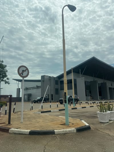 The image displays an exterior scene in Kwi, Nigeria, featuring a modern building and its immediate surroundings under a heavily overcast sky. The sky is filled with numerous small, uniform gray clouds, indicating a cloudy day with diffused lighting.

In the foreground, a paved area with an asphalt-like surface is visible. A prominent black and white painted curb defines the edge of a raised median or sidewalk, which is paved with light brown, rectangular pavers. Several black and white striped bollards or short posts are positioned along these paved areas.

A tall street lamp stands centrally, with a light-colored pole (beige transitioning to light blue-green at the base) supporting a dark gray, curved street light fixture. To the left, a standard circular "no U-turn" sign, white with a red border and a black U-turn arrow crossed by a red line, is mounted on a white pole. On the right, two white, ornate rectangular planters contain green snake plants (Sansevieria trifasciata).

The mid-ground features several parked dark-colored vehicles, including an SUV and a pickup truck, in what appears to be a parking lot or access road leading to the main building. A small, dark structure, possibly a security booth, is partially visible on the far left.

The dominant feature in the background is a large, multi-story, modern-style building constructed primarily with dark gray exterior materials. It has a prominent, steeply pitched dark roof overhang supported by visible struts. The building features multiple rectangular windows and several white security cameras mounted on its facade. A single, indistinct human figure is visible walking near the entrance on the right side of the building.