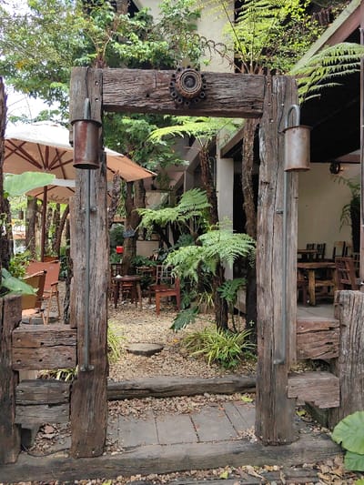 The image captures an unoccupied outdoor cafe or garden entrance located in Phú Xuân Ward, Vietnam. A prominent archway, constructed from four large, weathered dark timber beams, stands centrally. The archway features two vertical posts supporting a horizontal crossbeam, with a lower horizontal beam forming a threshold. A rusted, circular metal flange with visible bolt holes is affixed centrally to the top crossbeam. On the interior side of each vertical post, a rusted, cylindrical metal object, possibly a repurposed muffler or pipe, hangs from a curved metal rod.

Beyond the archway, a path of light-colored gravel contains a circular wooden stepping stone. To the left, several large, cream-colored patio umbrellas provide shade over multiple wooden tables and chairs. To the right, dense green foliage, including ferns and broad-leafed plants, surrounds additional wooden tables and chairs, positioned near a light-colored building with dark architectural elements partially visible. The immediate foreground shows ground covered with dark wooden planks, gravel, and scattered leaves. No individuals or active interactions are observed within the frame. The scene is illuminated by natural, diffused daylight.