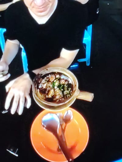 A person wearing a black t-shirt is seated at a dark table, with bright blue chair backs visible behind them. On the table, there is an orange plate with a spoon and fork resting on it. Next to the plate is a light-colored pot with a handle, filled with a dark-colored stew or soup topped with rice and green garnishes. The person's hands are visible, one reaching towards the pot, and the other resting on the table. The overall scene appears to be a close-up shot of a meal being served or eaten. There is no visual information to determine the location in Cherepovets, Russia.
