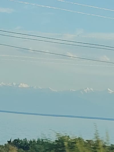 This image captures a serene natural landscape with a clear blue sky, a calm body of water, and a distant mountain range capped with snow. Three parallel power lines stretch horizontally across the sky, suggesting human infrastructure within the otherwise natural setting. The water appears to be a lake or a sea, reflecting the sky's light. Along the water's edge, there is a line of green foliage, possibly trees or bushes, with hints of drier brush in the foreground. The snow-capped mountains in the distance are a prominent feature, creating a sense of depth and grandeur. The overall impression is one of a peaceful, possibly early morning or late afternoon, with good weather. There are no people or specific objects of interest besides the natural elements and the power lines. The location context indicates this scene is in Bulan-Segettu, Kyrgyzstan.