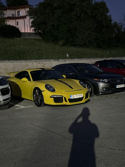 The image shows a yellow Porsche 911 GT3 parked in an asphalt parking lot. The car has black racing stripes on its hood and roof, and the license plate reads "CB 1919 PE". To the left of the Porsche is a white SUV, and behind it is a dark grey Honda Accord with the license plate "PB 9911 TA". Further back, a red car is partially visible. In the background, there is a building with a red roof and a lush green area with trees and grass. The scene appears to be in the city of Arbanassi, Bulgaria, as indicated by the building style and landscape. A shadow of a person holding a phone to take a selfie is cast on the asphalt in the foreground.