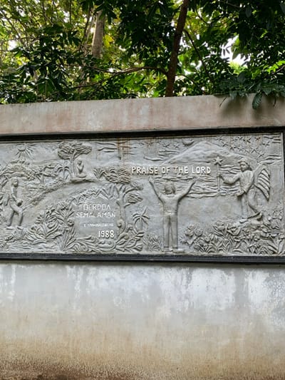 A gray concrete wall with a bas-relief sculpture depicting a scene of praise and worship. The sculpture is set within a black frame and features a man with his arms raised to the heavens. In the background, there is a mountain with a crescent moon in the sky, and another figure with wings holding a staff with a star on top. Below the sculpture are the words "Praise of the Lord," "Berdoa Semalaman," and "1 Tawarikh 28:10 1988" in white. The wall is situated in a forested area, with tree branches and leaves visible above it. The location is in Kecamatan Borobudur, Indonesia, based on the coordinates -7.606 latitude and 110.181 longitude.