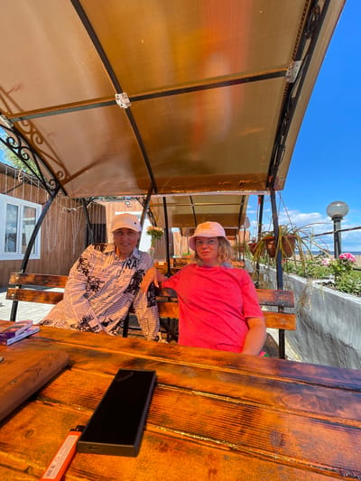 Two women are sitting on a wooden bench under a translucent amber-colored awning in an outdoor seating area. The woman on the left is wearing a white cap and a long-sleeved shirt with a newspaper print. The woman on the right is wearing a pink bucket hat and a bright pink t-shirt. They are seated at a wooden table. On the table are a mobile phone, a lighter, and a stack of cigarette packs. Behind them, there is a wooden building with string lights, potted plants, and a view of the sea and a beach in the distance. The sky is clear and blue with some clouds. This scene is located in Belozersk, Russia, likely at a cafe or restaurant with an outdoor seating area.