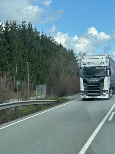 The image displays a daytime highway scene located in Kirchdorf am Inn, Germany, under a partially cloudy blue sky. A white Scania semi-truck, featuring a black grille and "SRT trans" branding, is prominently positioned on the right side of the asphalt road, traveling towards the viewer. The truck's front license plate, "A I-AF 752," is visible, indicating an Austrian registration. A silhouette of a driver is discernible within the cab. The road is marked with a solid white line on its right edge and double solid white lines separating traffic lanes. To the left of the road, a metal guardrail runs parallel to the pavement. Beyond the guardrail, the environment consists of deciduous trees and shrubs that appear leafless, backed by a dense line of evergreen conifers. A small, grey metal gate or fence structure is partially visible amidst the vegetation on the left side. The scene suggests early spring or late winter conditions.