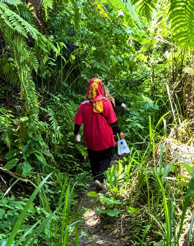 Two individuals are walking on a narrow dirt path through a dense forest. The person in the foreground is wearing a red t-shirt, black pants, and a headscarf with yellow and red patterns. They are holding a white plastic water jug in their right hand and a white bottle in their left. Another person, partially visible, is walking ahead of them. The path is surrounded by lush green vegetation, including ferns and tall grasses, with sunlight filtering through the leaves. The scene appears to be in Gunung Ambat, Indonesia, consistent with a jungle environment.