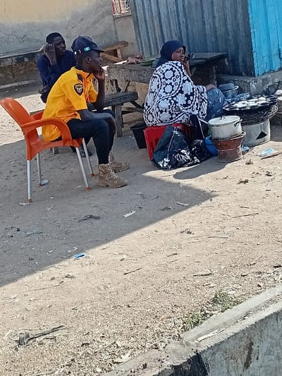 The image depicts an outdoor scene in Sharada, Nigeria. Three individuals are present. In the foreground, a male wearing a yellow collared shirt, dark trousers, a dark baseball cap, and tan tactical boots sits on an orange plastic chair, his right hand resting on his chin, looking toward the right. Behind him, a second male in a dark blue shirt is seated on a wooden bench, holding a dark object, possibly a phone, to his left ear. To the right, a female wearing a white and black patterned dress and a dark headscarf sits on a red plastic crate. She also holds a dark object to her left ear.
Adjacent to the female, a food preparation area is set up on the ground. This includes a metal charcoal stove with a round griddle on top, containing multiple light-colored items, possibly food. Several metal pots and containers, one stacked on another, are positioned near the stove. A black plastic bag is visible on the ground beside the red crate. The ground itself is dirt and sand, covered with scattered debris including wood chips and small pieces of paper. In the background, there is a building with a light-colored wall on the left and a corrugated metal wall, partly painted blue, on the right. A concrete curb or barrier runs along the lower right edge of the frame. Sunlight indicates daytime, casting distinct shadows across the ground.