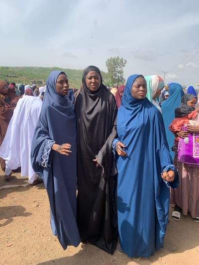 The image shows a group of women in Naraguta Hausawa, Nigeria. Three women are in the foreground, wearing different colored hijabs: blue, black, and blue again from bottom to top. These women are in line next to each other. The hands of the blue hijabs show henna and beaded embellishments. Other women can be seen in the background, wearing different colored hijabs and clothing. The ground is dirt.