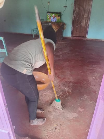 A person in black pants and a light shirt mops a concrete floor with a yellow-handled mop. A brown bucket sits nearby. Their bare feet are visible on the dirty floor. In the background, a TV shows an image above a wooden cabinet, positioned against a turquoise wall. A brown door is visible above the TV. A pink wall section is also visible.
