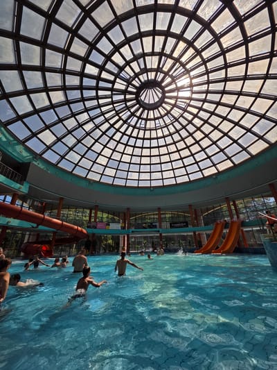 The image depicts an indoor swimming pool complex, likely located in Kobryn, Belarus, under a large, domed glass ceiling. The ceiling is segmented into a grid of square and diamond-shaped panes, allowing natural light to filter through, with the sun's glare visible in the center. The pool itself is filled with bright blue water, with several people swimming and playing. There are two orange water slides visible, one spiraling downwards and two others side-by-side. In the background, a lush green environment is visible through large windows, suggesting an outdoor setting beyond the indoor facility. Some signs are visible in the background, including one that appears to read "PROSTAPIZZA". The overall scene conveys a lively and recreational atmosphere within a well-lit, modern aquatic center.