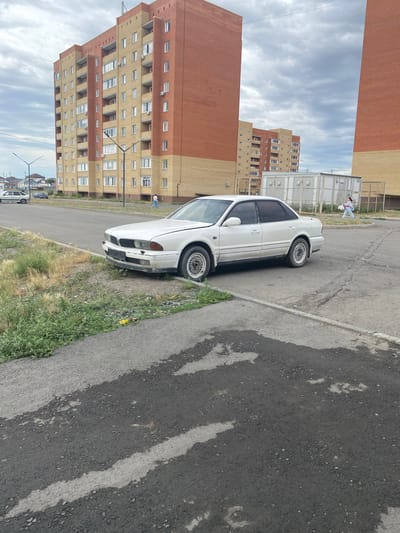 A white sedan is parked on the side of a paved road in Ekibastuz, Kazakhstan. The car, which appears to be older and possibly damaged, is positioned next to a grassy area and a sidewalk. In the background, there are multi-story apartment buildings with brick facades. One building is predominantly orange-red brick, while another is a lighter tan color. A person in light-colored clothing is walking in the distance, carrying a white object. The sky is overcast with clouds. The scene depicts an urban residential area.
