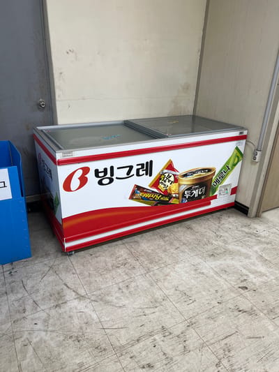 An ice cream freezer is shown against a textured, off-white wall and a gray tiled floor in Geoje-si, South Korea. The freezer has a white body with a prominent red stripe and an advertisement for Binggrae ice cream products. The top of the freezer consists of two transparent glass lids. Several ice cream products are depicted on the advertisement, including what appears to be a tub labeled "Together" and a green popsicle labeled "Melona." To the left of the freezer, a blue plastic bin is partially visible. The wall has some staining and a visible crack. A grey conduit runs vertically along the right side of the frame. The floor is worn and shows evidence of scuff marks and dirt.