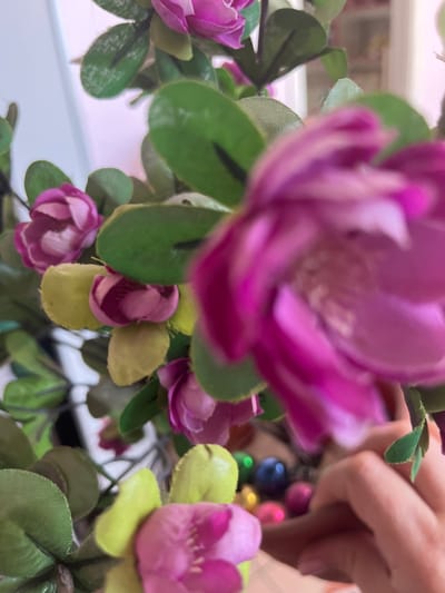 The scene depicts a close-up, shallow-depth-of-field view of artificial floral arrangements within an indoor setting in Кез, Russia. The foreground is dominated by a large, heavily blurred magenta-purple artificial flower, with its petals and a lighter center vaguely visible. Throughout the mid-ground, numerous smaller artificial flowers with magenta-purple petals and green fabric leaves are arranged on stems. Some leaves are a lighter, yellowish-green hue. Several of these smaller flowers appear partially open or in bud form. In the lower right background, a light-skinned human hand, possibly belonging to an adult, is visible, with fingers slightly curled, suggesting an interaction with objects beyond the immediate floral arrangement. Below the flowers and near the hand, a collection of small, reflective, spherical objects in various colors including blue, green, pink, and gold are observed, likely contained within a receptacle on a surface. The background is brightly lit and heavily blurred, consistent with an interior space.