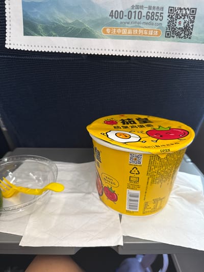 A close-up shot taken from a slightly elevated perspective shows a tray table on a train, likely in Qufu City, China, set up for a meal. The tray table is covered with a white paper napkin, and on it sits a bright yellow cup of instant noodles, with its lid partially open, revealing a cartoon egg and tomato. A clear plastic bowl containing a yellow fork and a piece of green vegetable is to the left of the noodles. Behind the tray table, dark blue upholstery of a train seat is visible. At the top of the image, a promotional banner with Chinese text, a phone number "400-010-6855", a website address "www.rimai-media.com", and a QR code can be seen. The lighting suggests an indoor setting, possibly during the daytime given the ambient light illuminating the scene.