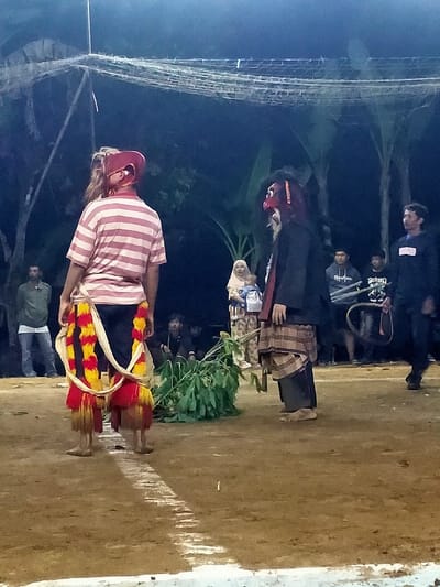 This image depicts an outdoor night scene in Tamansari, Indonesia. The ground is packed earth, marked with a white painted line in the foreground that extends towards the midground. Two central figures stand in the midground, facing each other.
The figure on the left is positioned with their back partially to the viewer, wearing a horizontal striped pink and white short-sleeved shirt over dark trousers. Their head is covered by a red, mask-like headpiece concealing the eyes, from which light-colored hair extends down the back. Red and yellow fringed sashes are wrapped around their waist and legs. This individual is barefoot.
The figure on the right stands facing the first figure. They wear a dark long-sleeved top, a brown and tan plaid sarong wrapped over dark trousers, and a dark red mask featuring a prominent nose and dark, fibrous attachments resembling hair. This individual is also barefoot.
Between these two figures, a pile of green leafy branches lies on the ground. In the background, several individuals are observing the scene. To the far left, a man in a light green long-sleeved shirt stands. To the center-left, a woman in a light-colored hijab holds an infant. Further right in the background, multiple men are present, some standing, one holding a large dark hoop and other objects. Overhead, a light-colored net or mesh structure is visible against the dark background, which consists of trees and dense foliage. The scene is illuminated by an unseen artificial light source, suggesting an evening or night event.