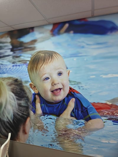 An indoor scene in Warrington, United Kingdom, featuring a large, high-resolution wall mural. The mural depicts a Caucasian infant, estimated age 6-18 months, with light hair and blue eyes, smiling broadly while partially submerged in blue water, indicative of a swimming pool. The infant wears a blue and red short-sleeved swimming top and is supported by two adult hands, also depicted within the mural. Water droplets are visible on the infant's face and hair. In the immediate foreground, the back of an adult's head and left shoulder, with light-colored hair tied back, is visible, positioned to observe the mural. Above the mural, a white suspended acoustic ceiling with a grid pattern is present. A light-colored horizontal surface or partition runs across the bottom of the frame. A fragment of a white furniture item is partially visible in the extreme bottom-left corner. The specific venue within Warrington is not identifiable from the visual data.