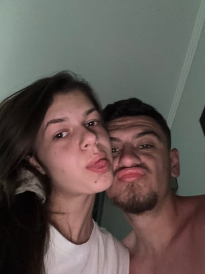 The image is a close-up, high-angle selfie featuring two young individuals, a male and a female, looking up towards the camera. The male, positioned on the upper right side of the frame, has dark hair and light facial stubble. He is making a humorous puckered or "fish" face with his lips. He appears to be shirtless or wearing a very light-colored garment, with his shoulder and chest visible.
The female, situated below and to the left of the male, has long, dark hair and is playfully sticking out her tongue. She is wearing a white top. Both subjects display a casual and affectionate demeanor.
The setting appears to be indoors, with a plain, light-colored wall or ceiling forming the background. A subtle architectural detail, possibly a crown molding or trim, is visible in the upper left corner of the frame. The lighting is dim, suggesting it might be evening or night, or simply an indoor environment with low light. There are no visible texts or specific location cues in the image.