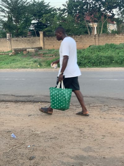 A man is walking along the side of a paved road in Gbeleko, Nigeria. The man is wearing a white shirt, black shorts, and sandals. He is carrying a green bag with white polka dots in his right hand and a white object in his left. The road is cracked and has some debris on the side. On one side of the road is green vegetation and a partially constructed building made of cinder blocks. On the other side is a dirt shoulder with some dry vegetation. The sky is overcast.