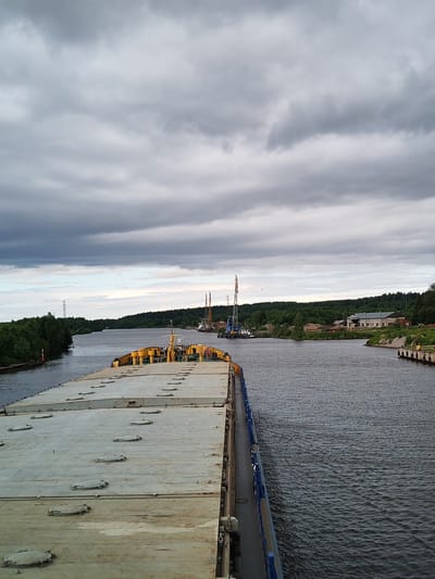 This is a medium shot taken from a vessel moving on a calm body of water under an overcast sky. The foreground is dominated by the deck of the vessel, which is flat and covered with numerous circular hatches. The vessel has a yellow superstructure visible on the right side of the deck. The water is dark and shows gentle ripples.
In the mid-ground, the waterway widens, flanked by lush green trees and vegetation on both banks. On the left bank, there's a tall transmission tower and a red and white marker buoy in the water. On the right bank, a complex of industrial buildings with warehouses and a dock with bollards is visible. In the distance, on the water, two large crane barges are positioned, suggesting construction or industrial activity. The sky is filled with heavy, gray clouds, creating a subdued and somewhat dramatic atmosphere. The scene appears to be set in a canal or river, possibly part of the Vyshny Volochyok Water System or the White Sea–Baltic Canal, given the industrial infrastructure and the presence of barges, which is consistent with locations near Vytegra, Russia.