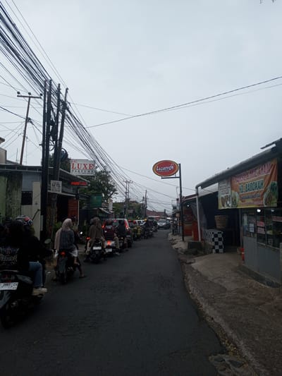 The image captures a congested street scene in the city of Houaythat, Laos, under an overcast sky. An asphalt road, appearing worn, is filled with stationary traffic, predominantly motorcycles and scooters, with a few cars visible further down the street. Multiple individuals are seated on the motorcycles, some wearing helmets, others with head coverings.
On the left side of the street, a building houses commercial establishments. Prominent signage, primarily in Indonesian, includes "LUXE PARFUME" and "ALAT VITAL" with a phone number "0821-2290-0424." A dense array of black utility wires crisscrosses overhead, supported by several utility poles.
On the right side of the street, a red and yellow sign for "Lazatto Chicken & Burger" is affixed to a pole. Adjacent to it, a large banner, also in Indonesian, advertises "BAKSO & MIE AYAM HD. BAROKAH," featuring images of food items. The ground near the sidewalk on the right shows uneven concrete and a low wall or counter with a black and white checkered pattern.