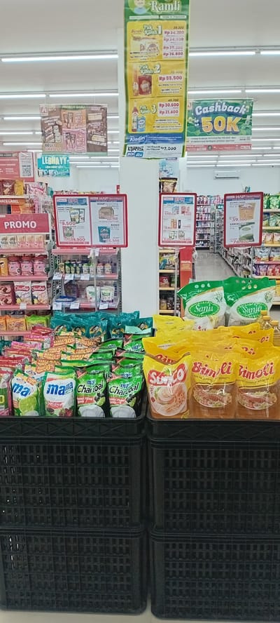 The image shows an aisle in a supermarket in Payacumbuh, Indonesia. The shelves are stocked with various food items, including cooking oil, instant noodles, snacks, and condiments. Promotional banners advertise cashback offers and special deals on specific products. Black plastic crates are visible on the right side of the frame. The lighting is bright and fluorescent. No people are visible in the frame.