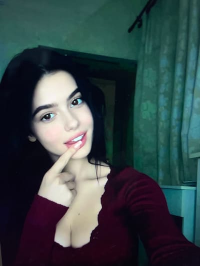 The image shows a close-up of a young woman with dark hair and brown eyes, wearing a burgundy top. She is holding a finger to her lips and looking at the camera with a slight smile. The background appears to be indoors, with a curtain and a doorway visible. The lighting is dim, casting a green hue over the scene. There is no visible indication of the location being the city of Belyye Berega, Russia.