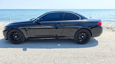 This media file showcases a black BMW convertible parked on a gravel surface near a body of water, likely the sea. The car is the primary subject, viewed from its side profile. It features tinted windows and black rims, with a red taillight visible on the rear.

The background reveals a calm, blue sea stretching to the horizon under a clear, bright sky, suggesting a sunny day. The shoreline to the left is composed of sand and pebbles, with a rustic wooden post partially submerged in the water. To the right, the shore transitions to a rocky beach. There are two small, dark figures visible in the water in the distance, possibly swimmers or buoys.

The overall impression is one of a peaceful, coastal setting on a warm, clear day. No people are clearly visible interacting with the car or the environment. The lighting indicates it is daytime, likely late morning or early afternoon given the bright sun and clear visibility. The location context of Leptokarya, Greece, aligns with the coastal scenery. There is no visible text in the image.