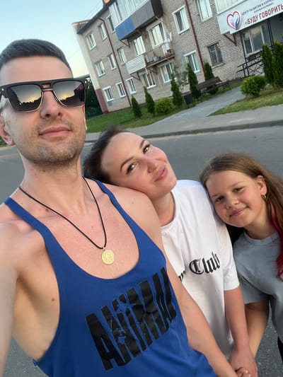 The image shows a family of three taking a selfie outdoors. The subjects are two adults and a child, likely a father, mother, and daughter. The father is in the foreground, wearing sunglasses and a blue tank top with a black graphic. He has short, dark hair and stubble. The mother is lying down behind him, looking at the camera with a gentle smile. She has dark hair and is wearing a white t-shirt. The daughter is resting her head on the mother's shoulder, also looking at the camera with a bright smile. She has long, brown hair with streaks of pink and is wearing a light blue t-shirt.

The scene appears to be in a semi-urban environment. Behind the people, a building with brickwork and multiple windows is visible, along with some greenery. The ground seems to be paved, possibly a sidewalk or street. The lighting suggests it's daytime, likely late afternoon or early evening, with soft light casting reflections in the father's sunglasses. The overall mood is happy and relaxed, reflecting a moment of family connection.

A sign with text is partially visible on the building, but it is too blurry to read clearly. The graphic on the father's tank top is also difficult to decipher. The daughter's t-shirt has the word "Club" in gothic-style font. There is a small, circular, golden pendant on a black string around the father's neck.