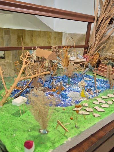 This image depicts a diorama representing a wetland ecosystem. Key elements include a body of water simulated with blue materials, green "grass" terrain dotted with model trees and vegetation. Model birds, specifically pelicans and other waterbirds, are placed in and around the water. A small, constructed structure (possibly a hut or bridge) is also present. Dried plants and branches are used for additional landscaping effects.
While not explicitly stated in the image, given the prompt, the diorama is most likely intended to represent the Srebarna Nature Reserve in Bulgaria, a UNESCO World Heritage Site known for its diverse birdlife.