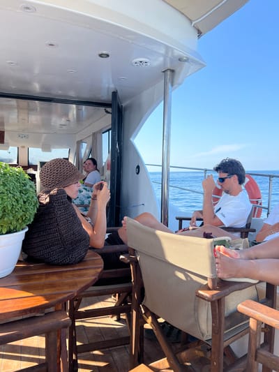 On a sunny day, a group of people relax on the deck of a boat on the Aegean Sea, near the coast of Skopelos, Greece. The boat appears to be a small yacht or ferry with a white superstructure and a wooden deck. In the foreground, a woman wearing a brown crocheted hat and a dark knitted garment is seated at a wooden table, holding a smartphone in her hands. To her right, a man in a white t-shirt and sunglasses is seated, looking out at the sea. A life preserver is visible on the railing behind him. Other passengers are seated in chairs, some with their legs propped up. The water is calm and blue, and the sky is clear. Green foliage is visible in a white pot on the left side of the image.