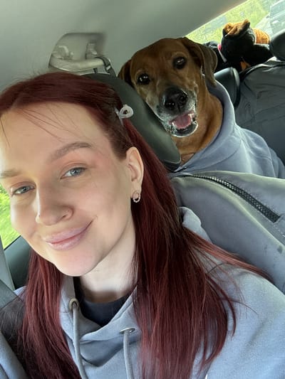 A selfie photograph shows a woman and a dog in the interior of a vehicle. The woman has light skin, red hair, and is wearing an earring. A clear hair clip is visible in her hair. She is looking at the camera and smiling.
The dog is medium-sized with brown fur. It has its mouth open, appearing to smile. The dog is wearing a gray hoodie.
The scene appears to be inside a car. The background shows parts of the car's interior, including a headrest, window, and seat. The location in the city of Balakhna, Russia cannot be determined from the visual information in the photograph.