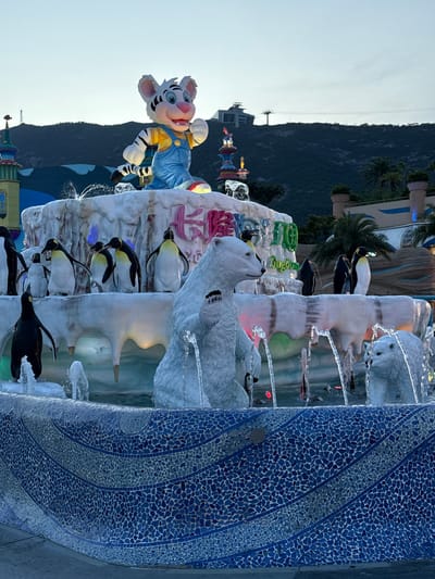 A water fountain sculpture featuring polar bears and penguins is prominently featured against a backdrop of mountains. A cartoonish tiger figure wearing overalls stands to the left. The fountain is constructed with white, icy-looking material, with flowing water. A blue mosaic column stands to the right. This is located in Xiangzhou, China.
