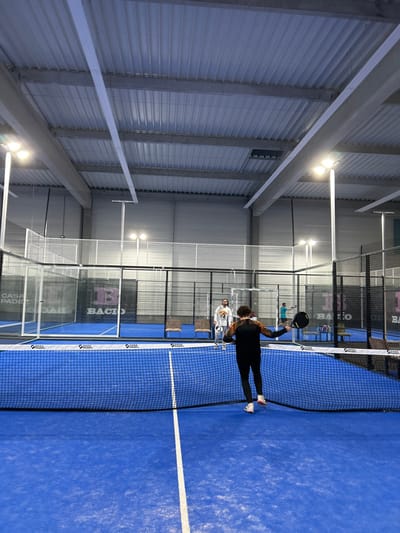 A woman in black athletic wear and white sneakers stands on a blue padel court. She holds a black padel racket overhead as if in the middle of a swing. A white line runs across the court. Behind her, another person in white stands near seating. The court is enclosed with a net and glass walls bearing logos. Above, the ceiling is corrugated metal with rows of fluorescent lights.