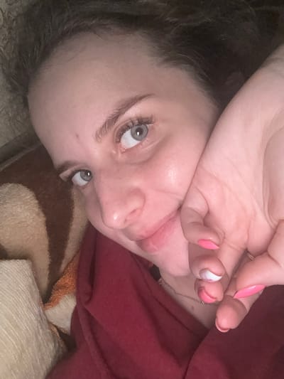 The media file contains a close-up, tilted shot of a person's face and hand, with a blanket and red fabric in the background. The person has fair skin, light blue eyes, and dark hair. Their left hand is raised to their face, partially covering their forehead and eye. The person's lips are slightly parted, and their tongue is visible. They are wearing a red garment and lying on a striped blanket. The nails on the visible fingers are painted pink and white. The lighting appears to be dim, suggesting an indoor setting, possibly at night or in a dimly lit room. The overall mood of the image is intimate and somewhat suggestive, due to the pose and facial expression. There is no visible text in the image.