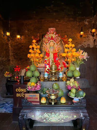 The image depicts an altar inside a dimly lit brick structure, likely a temple or shrine, located in Tuy Hoa Ward, Vietnam. The central focus is a richly adorned statue of a deity, possibly a goddess, wearing a red and gold robe and an elaborate headdress. Flanking the statue are tall, golden floral arrangements and a bouquet of pink and white lilies in a vase. In front of the statue, on a multi-tiered offering table, are various fruits including pomelos, dragon fruits, oranges, and a watermelon. Incense sticks in a brass urn and small candle holders with yellow flames are also present. To the left of the altar, a dark wooden cabinet displays more fruits and what appear to be red flowers in a vase. Two yellow lanterns hang on the brick walls, providing ambient light. A sign with Vietnamese text, "CÔNG ĐỨC THIÊN Y A NA," is visible on a dark wooden box in the foreground. The overall atmosphere is one of religious reverence and offering.