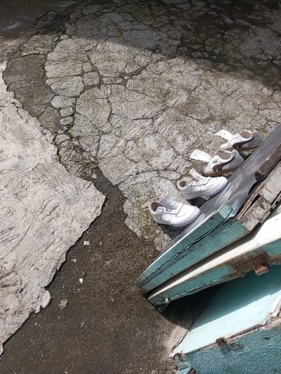 Cracked concrete meets dark asphalt. Four pairs of shoes, arranged in a line, are positioned on the concrete. Three pairs are light brown and white, while the pair closest to the corner is all white. The shoes are slightly angled and rest against a stack of light blue wooden boards with rusty hinges. The scene is outside and lit by sunlight.