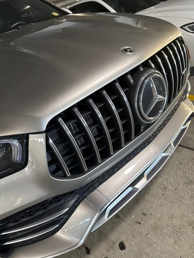 A close-up shot shows the front of a silver Mercedes-Benz, specifically the grill with the iconic Mercedes-Benz star emblem prominently displayed in the center. The grill features horizontal chrome bars. A smaller Mercedes-Benz emblem is also visible on the body of the car to the left of the grill. Part of the headlight assembly is visible at the bottom of the image. The car is parked in an enclosed area, possibly a garage or parking structure, with a concrete wall visible in the background. The location is Hollywood Seminole Reservation, United States of America.